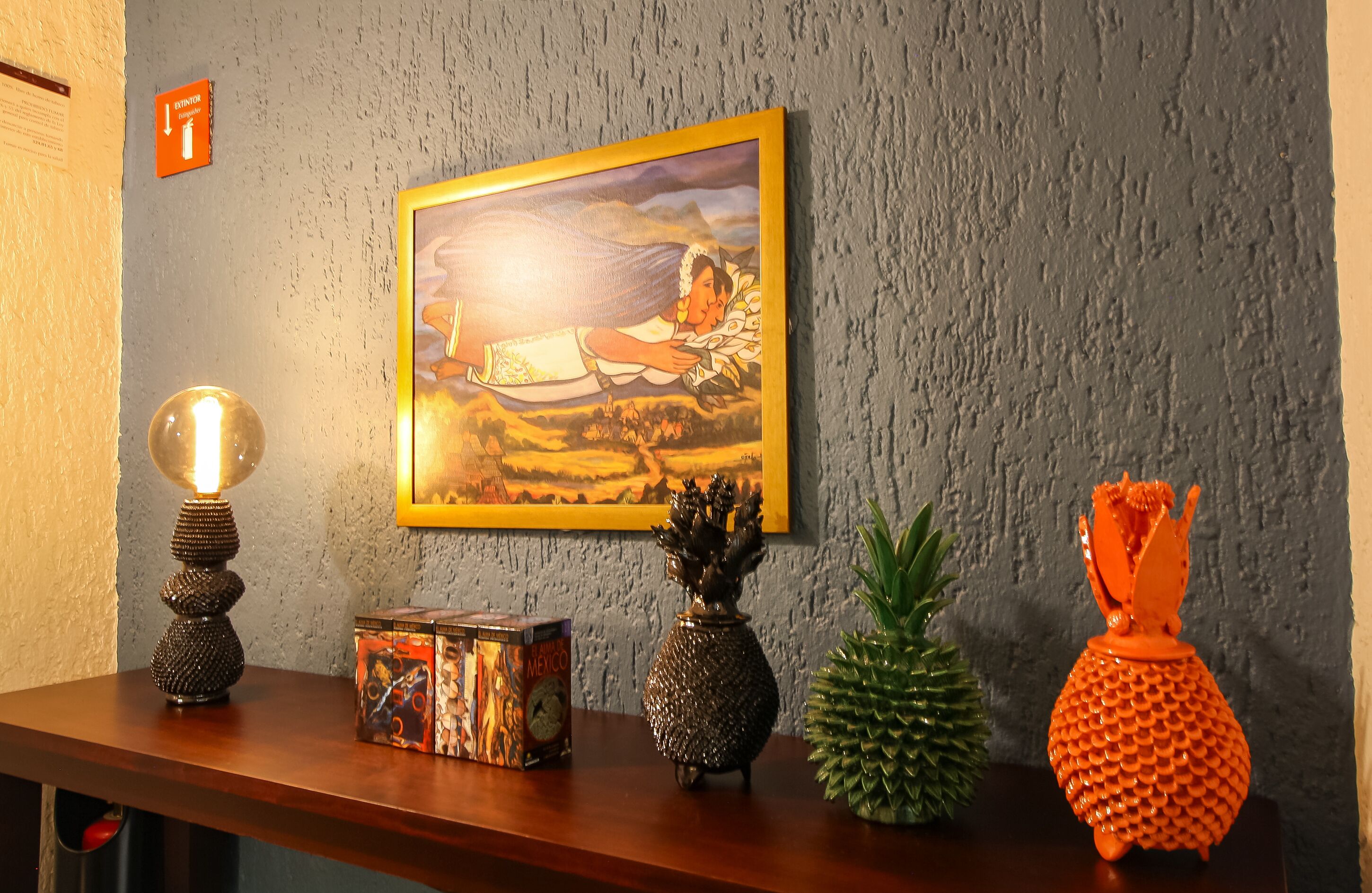 Una pared con un cuadro enmarcado, una lámpara de lava, una caja de juegos de mesa con temática de México, un florero con forma de piña y una planta de piña.