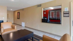 Un hall de hotel con paredes color beige, un espejo grande, una recepción y una zona de estar con dos sillas y una mesa.