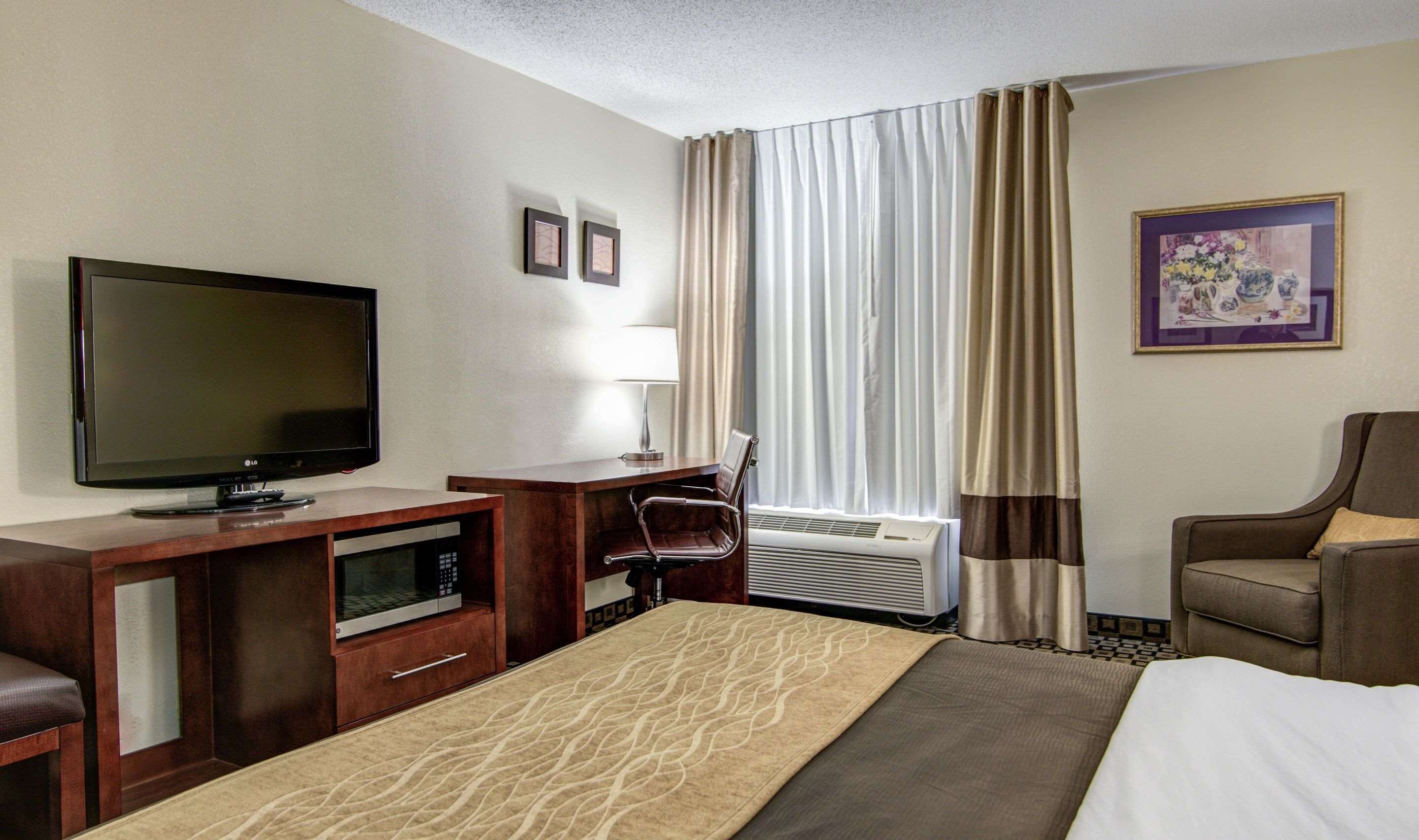 A hotel room with a bed, a desk with a flat-screen TV, a chair, a microwave, and a window with curtains.