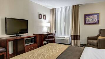 A hotel room with a bed, a desk with a flat-screen TV, a chair, a microwave, and a window with curtains.