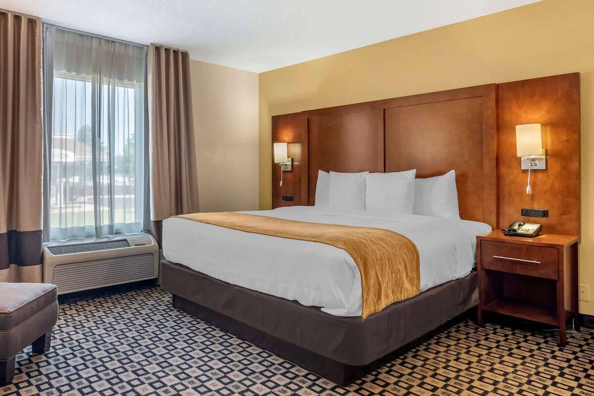 A hotel room with a large bed, a bedside table with a phone, a chair, and a window with curtains.