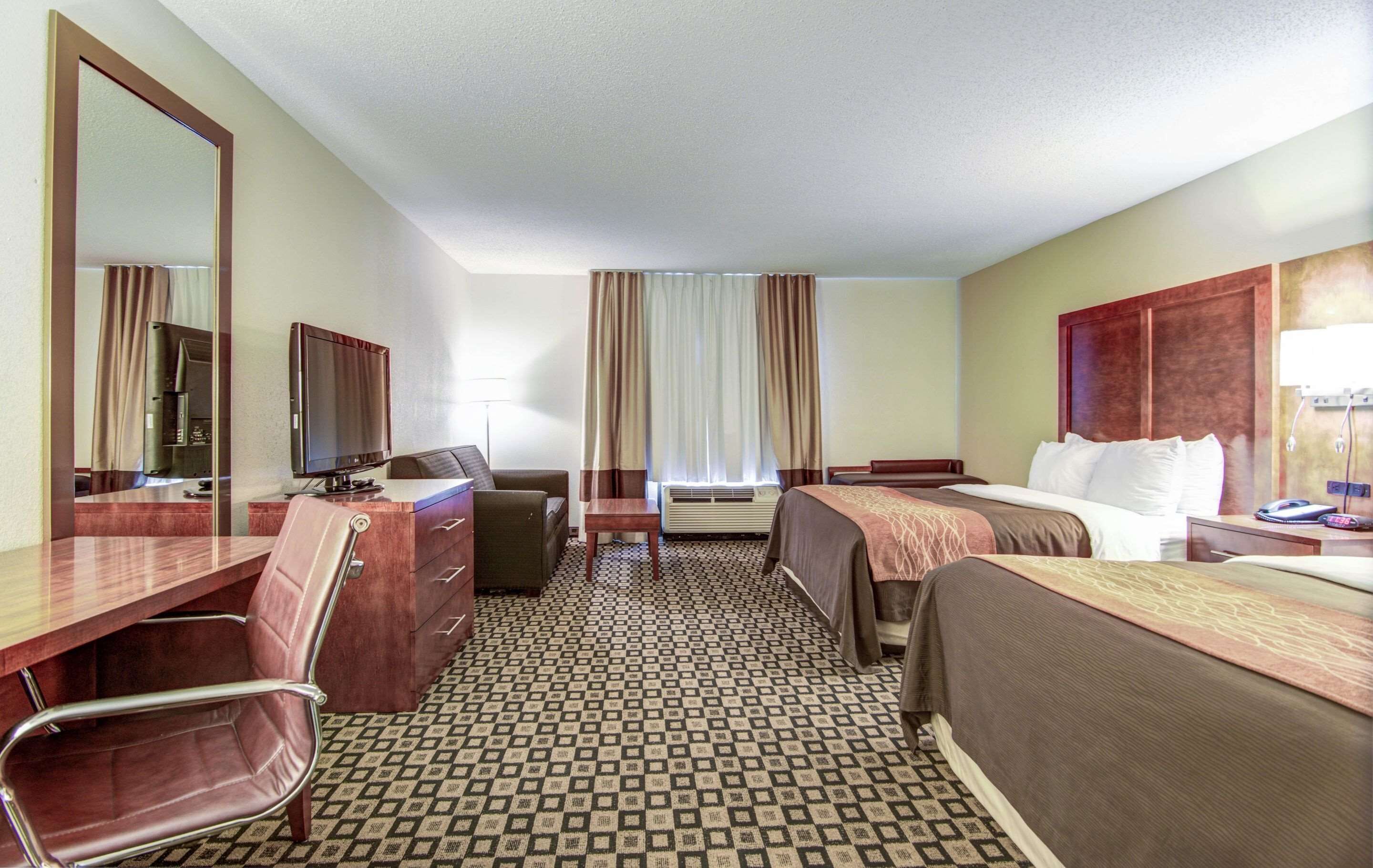 A hotel room with a desk, two beds, a chair, a TV, and a large mirror.