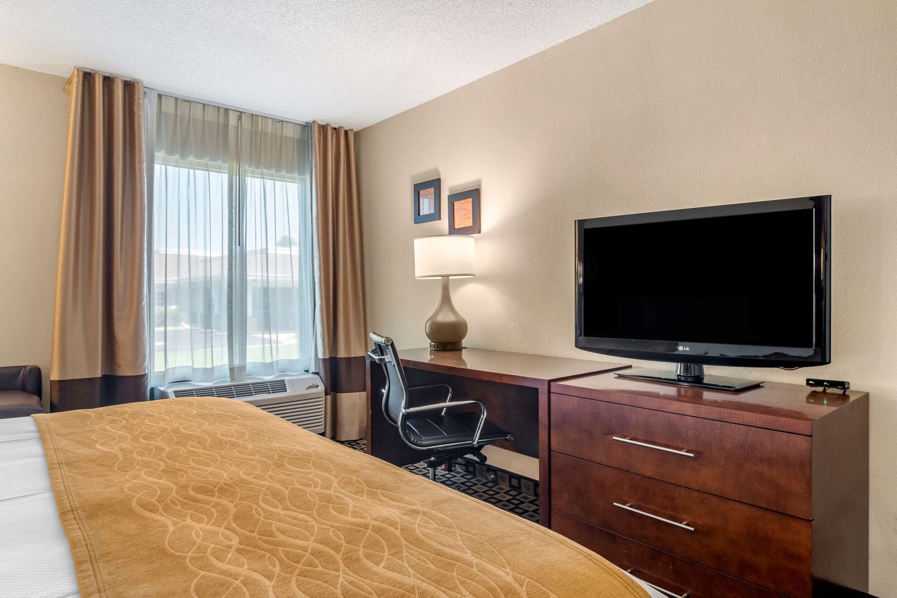 A hotel room with a bed, a desk with a television, a chair, a lamp, and a window with curtains.