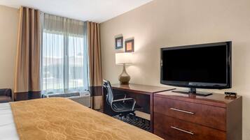 A hotel room with a bed, a desk with a television, a chair, a lamp, and a window with curtains.