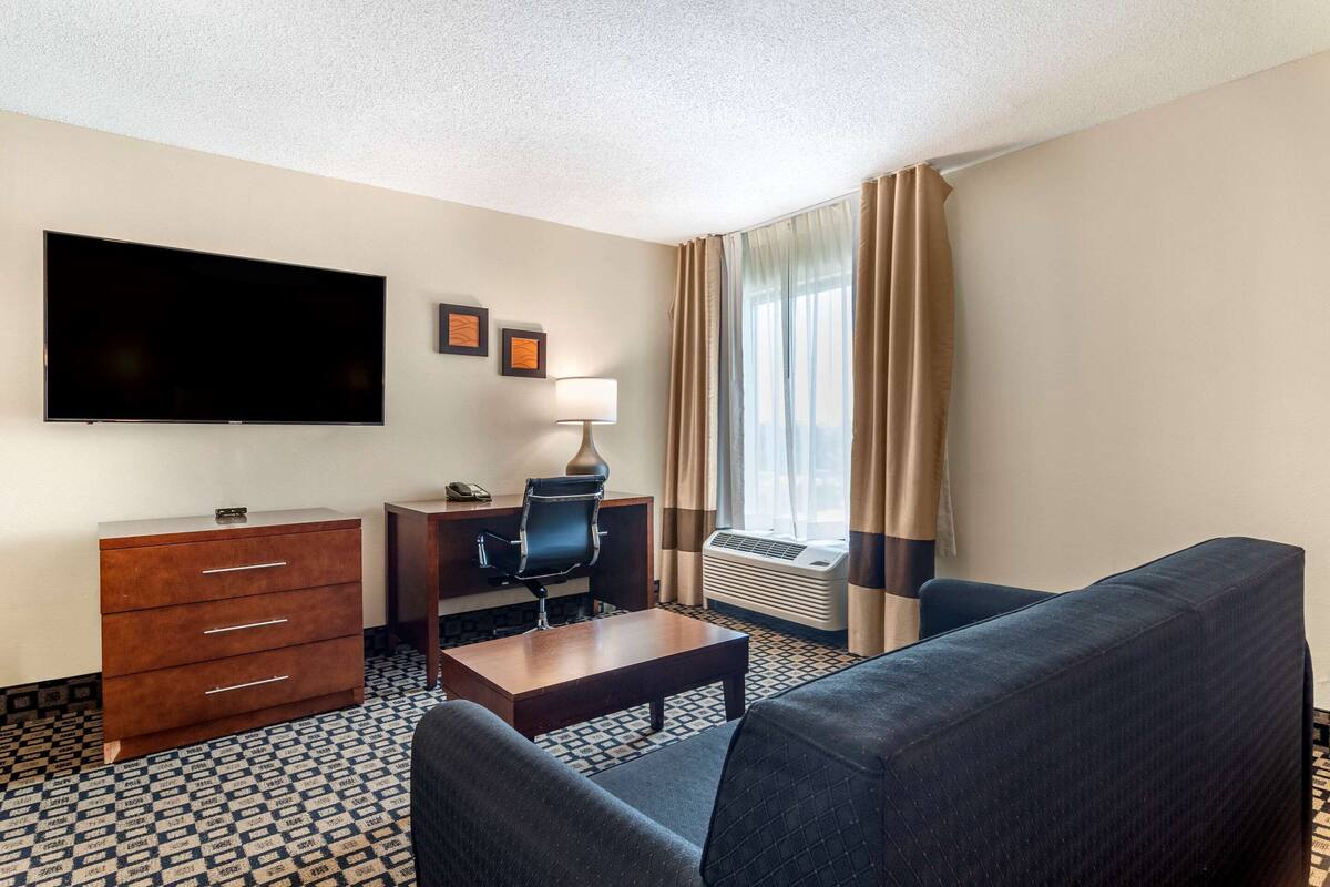 A hotel room with a flat-screen TV, a desk with a chair, a sofa, a dresser, and a window with curtains.