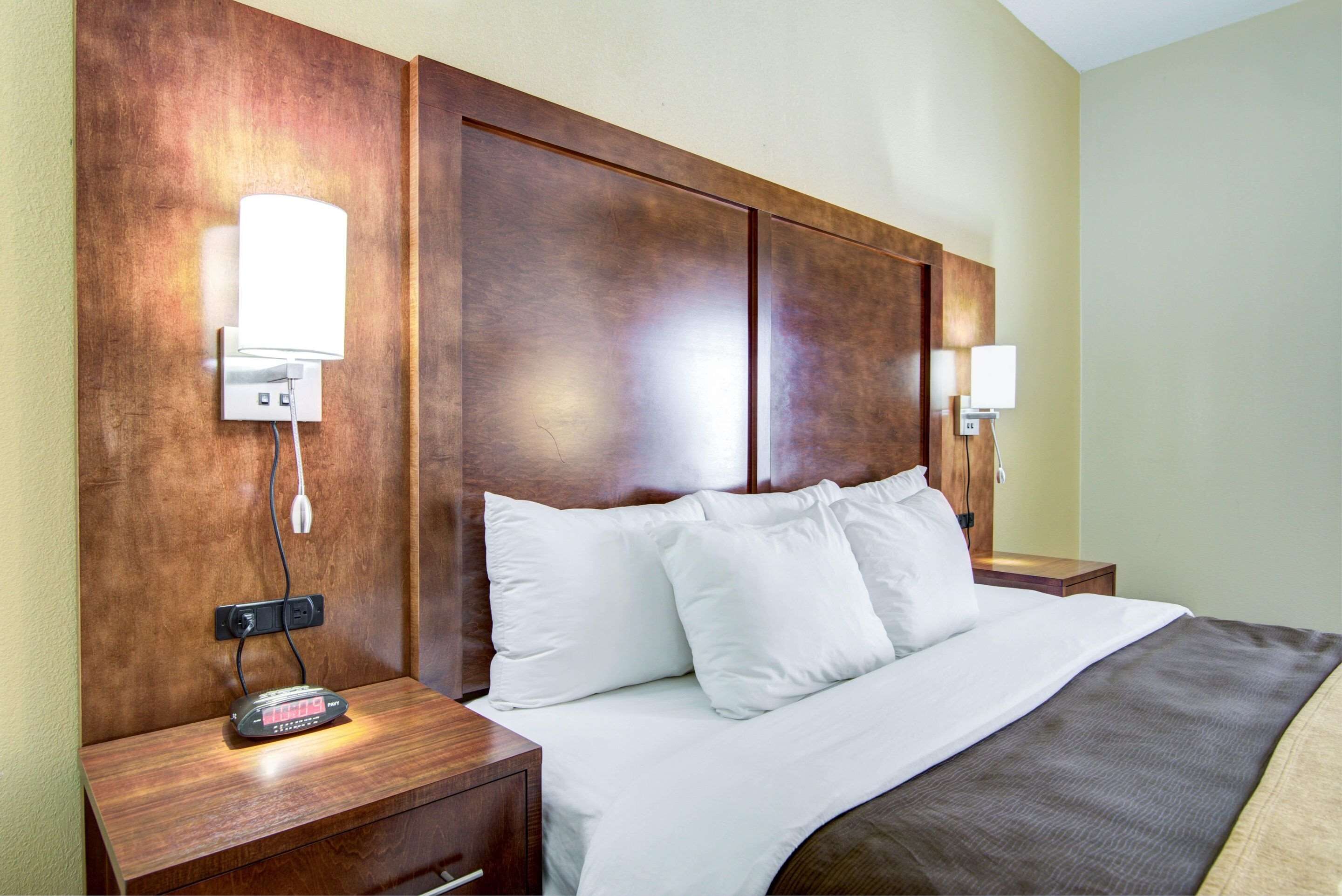 Quarto de hotel com cabeceira de madeira, roupa de cama branca, mesa de cabeceira com abajur e régua de tomada.
