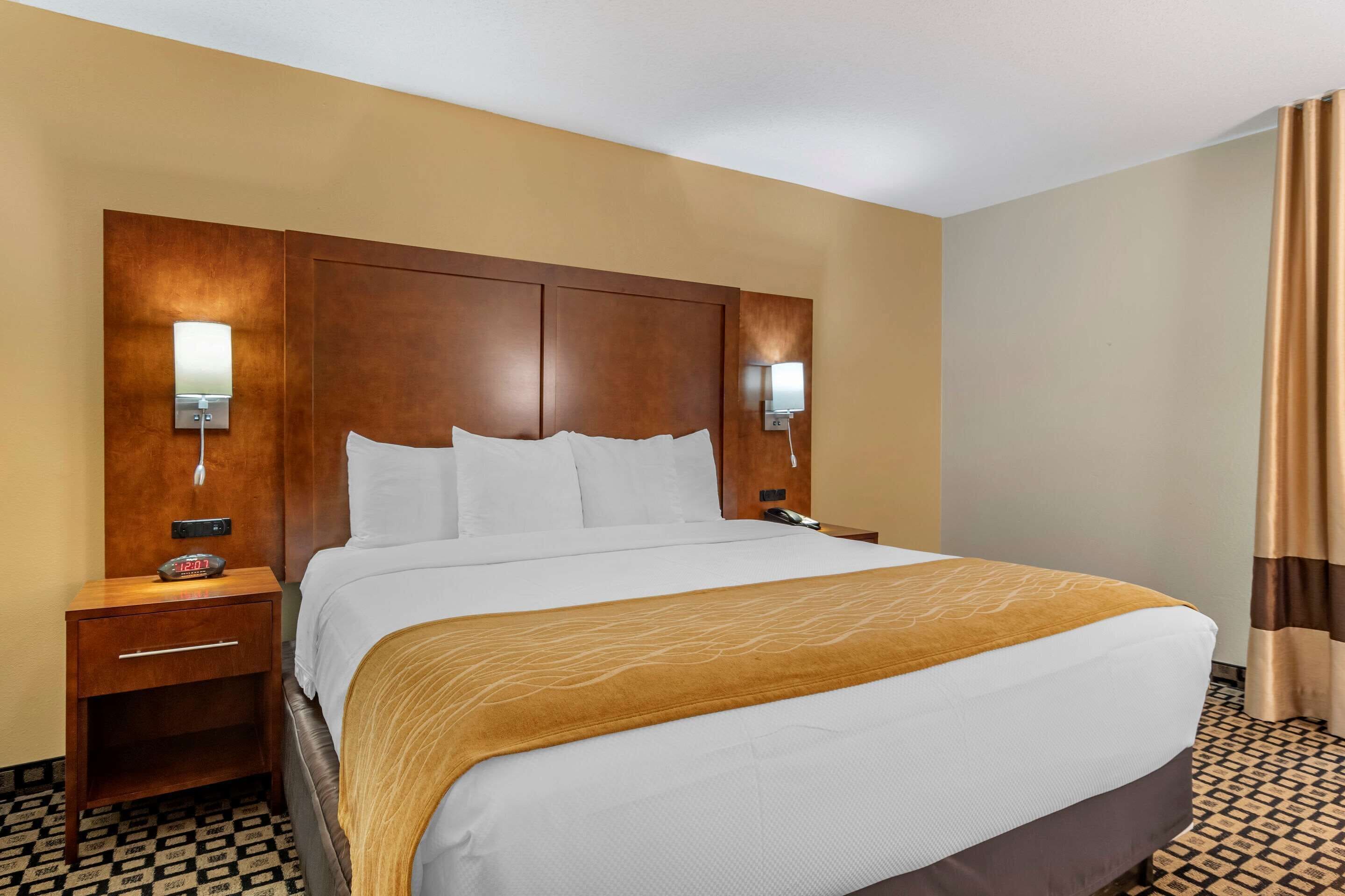 A hotel room with a large bed, wooden headboard, bedside lamps, a nightstand with a clock, and patterned floor tiles.