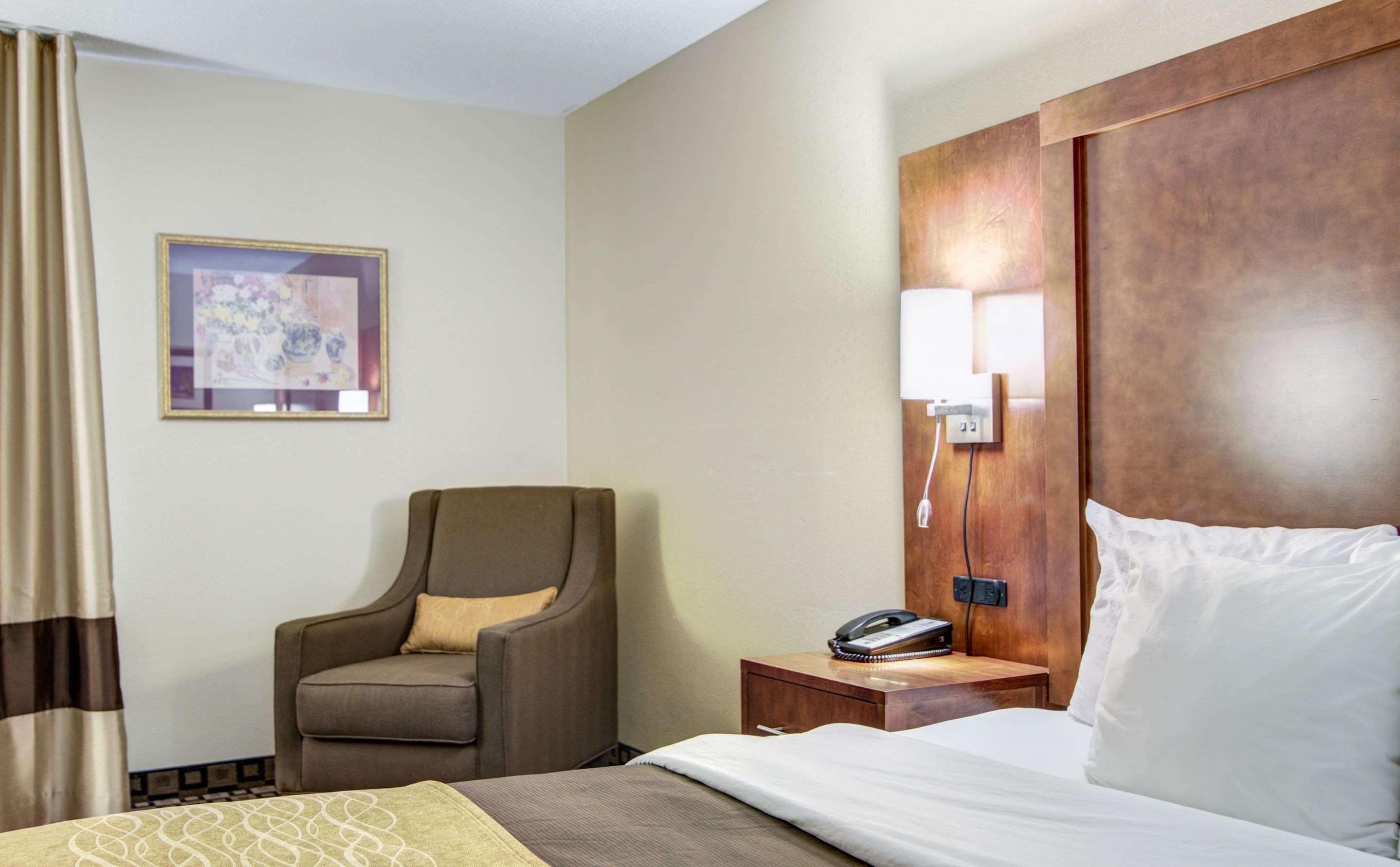 A hotel room with a bed, a chair, a nightstand with a phone, a wall-mounted lamp, and a framed picture on the wall.