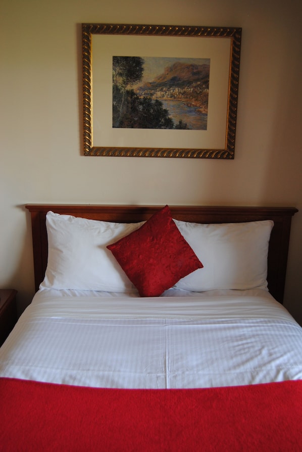 A neatly made bed with white linens and a red accent pillow, a framed landscape painting above it, and a wooden headboard. - Economy Inn (Edmonton)