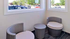 A corner of a room with two wicker chairs and a small round wicker stool, a white toilet, and a view of parked cars through a window.
