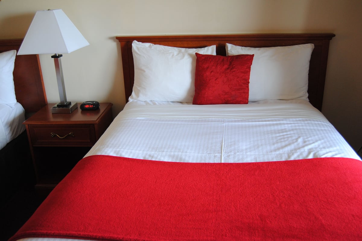A hotel room with a neatly made bed, a bedside table with a lamp and alarm clock, and a red throw blanket.
