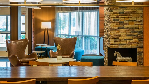 Un vestíbulo de hotel con chimenea de piedra, sillones de cuero, una mesa redonda y vistas al exterior a través de amplios ventanales.