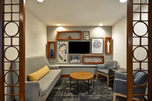 Uma sala de estar com um sofá cinzento, uma mesa de centro redonda de madeira, e duas poltronas. Há quadros nas paredes, uma televisão de tela plana e uma estante de madeira decorativa.