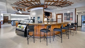 Zona de bar moderna con una barra central, varios taburetes y un área de asientos con techo alto y vigas de madera.