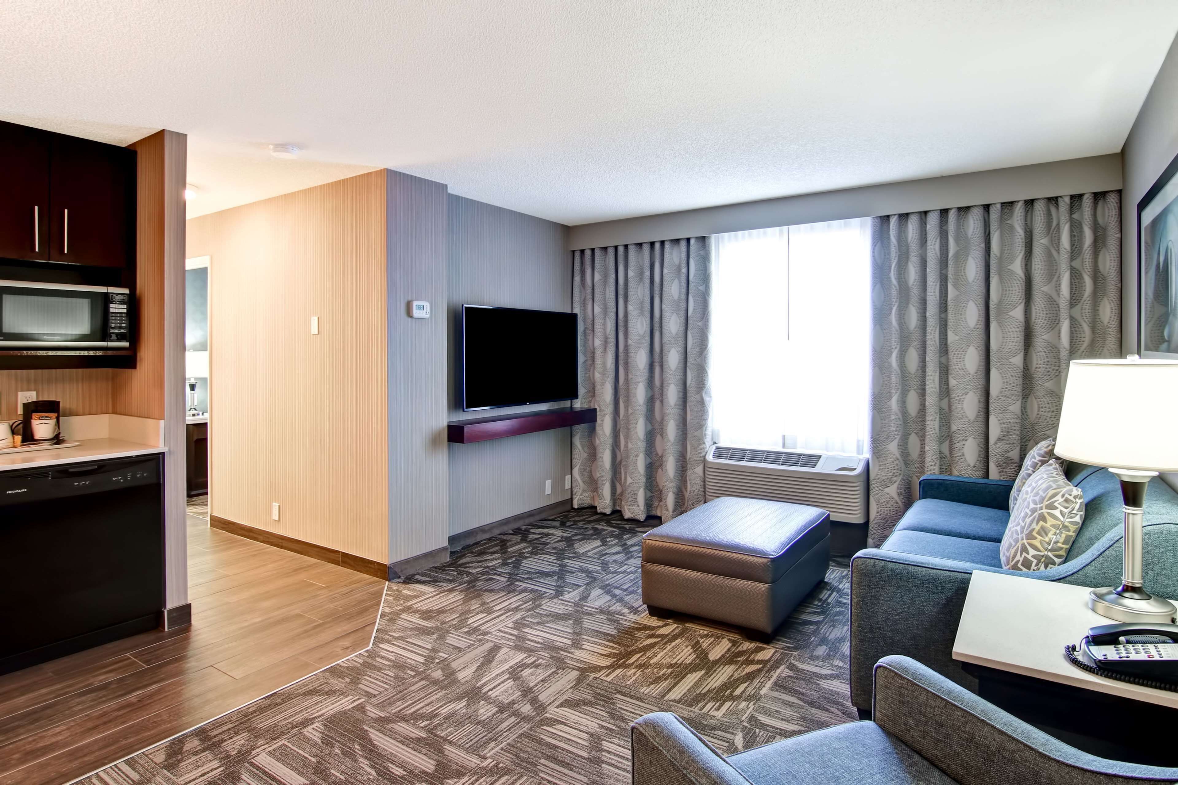 Ett hotellrum med ett litet kök, en platt-TV, en soffa och ett fönster med gardiner.