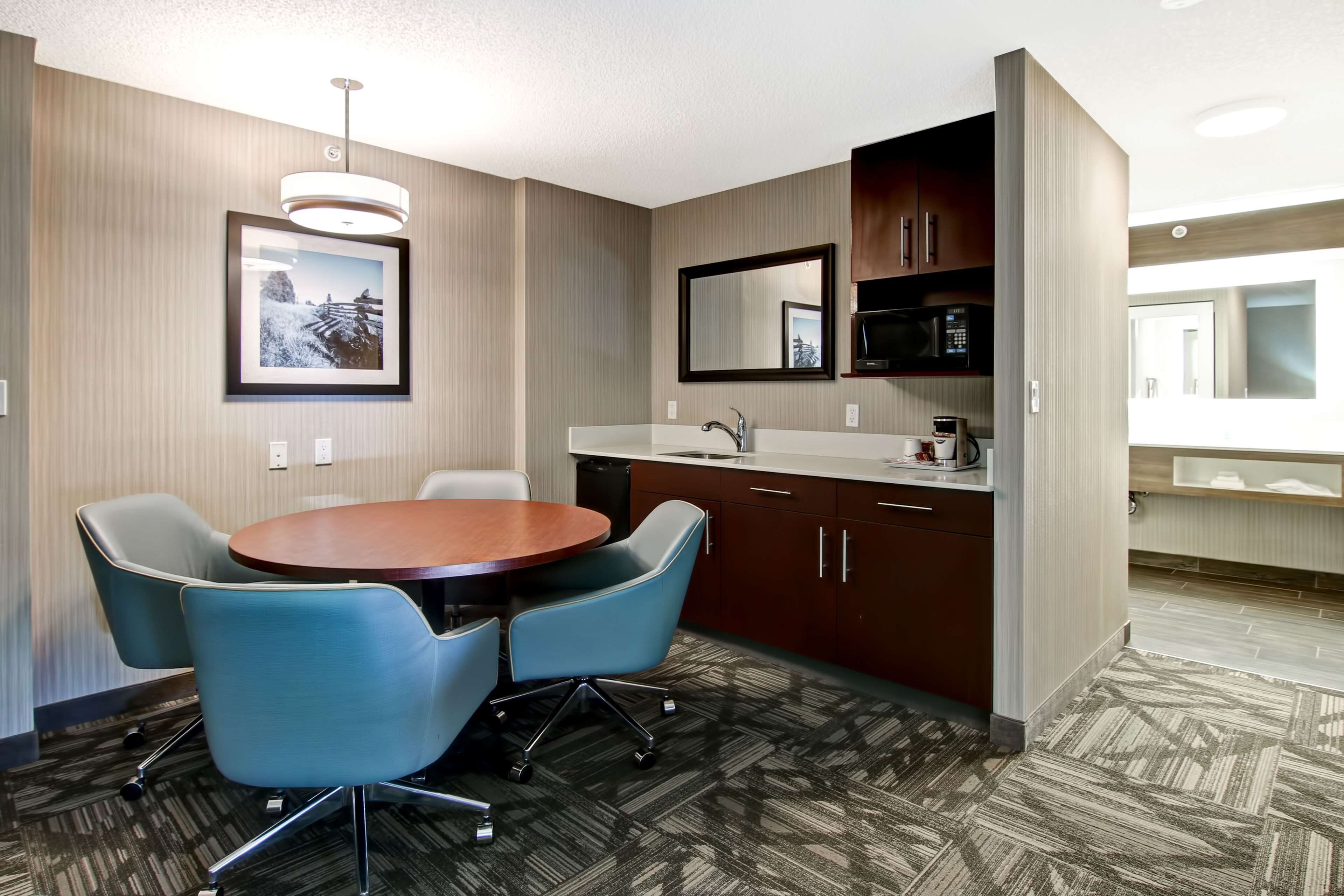Una habitación de hotel moderna con una mesa de comedor redonda, dos sillas azules, una cocina integrada con microondas y un espejo grande sobre el lavamanos.