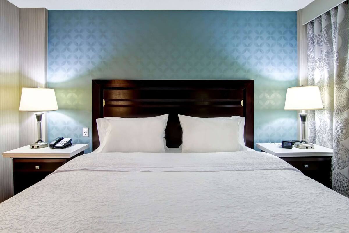 A hotel room with a large bed, two bedside tables, a nightstand, a wall clock, and a patterned wall.