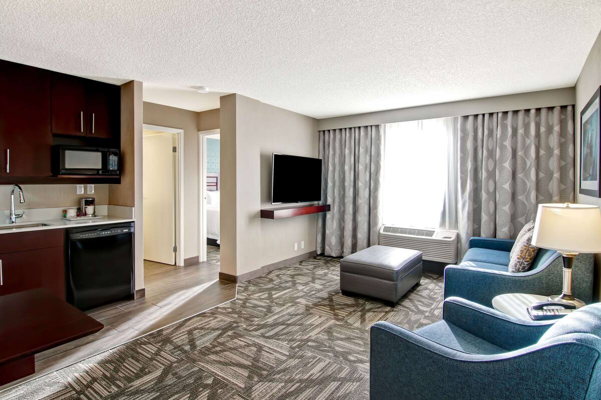 A hotel room with a kitchenette, a flat-screen TV mounted on the wall, a seating area with a sofa and ottoman, and a large window with patterned curtains.