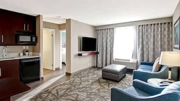 A hotel room with a kitchenette, a flat-screen TV mounted on the wall, a seating area with a sofa and ottoman, and a large window with patterned curtains.