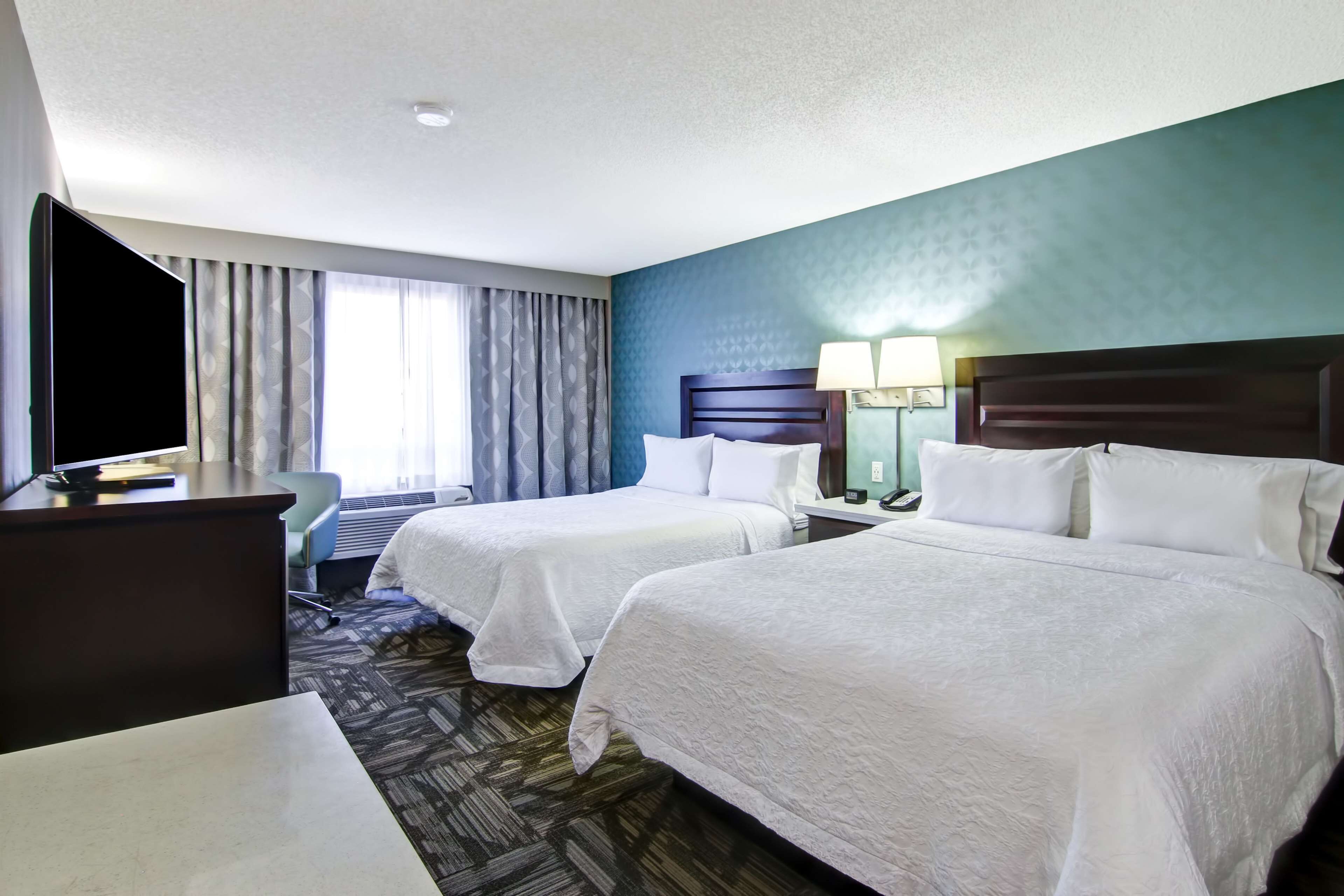 A hotel room with two beds, a desk, a TV, and a window with curtains.
