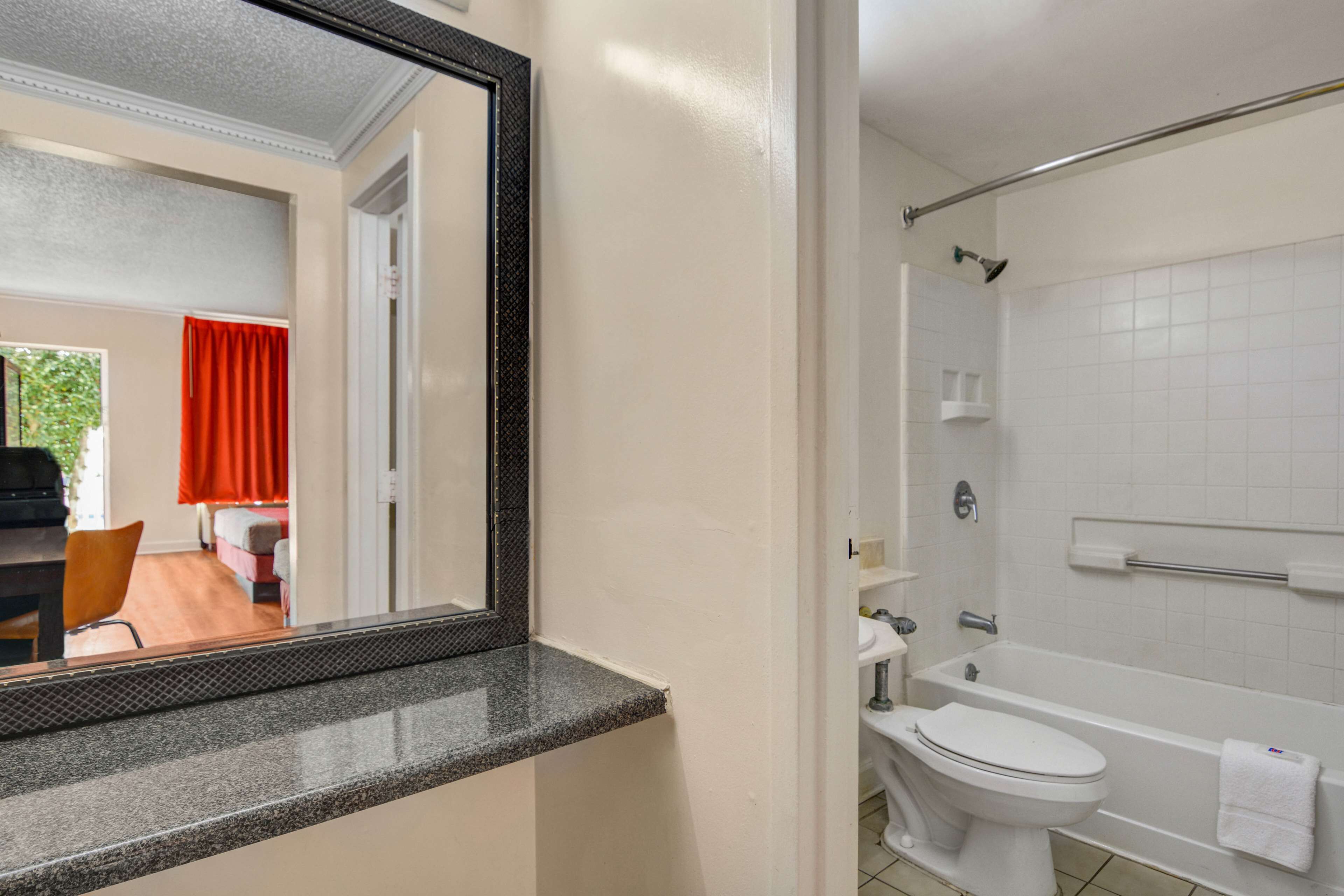 Une salle de bain avec des toilettes, un lavabo et une baignoire, tous de couleur blanche. Un miroir est placé au-dessus du lavabo, et une tringle à rideau de douche se trouve au-dessus de la baignoire.
