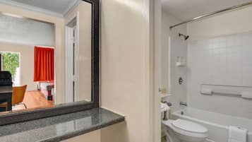 Une salle de bain avec des toilettes, un lavabo et une baignoire, tous de couleur blanche. Un miroir est placé au-dessus du lavabo, et une tringle à rideau de douche se trouve au-dessus de la baignoire.