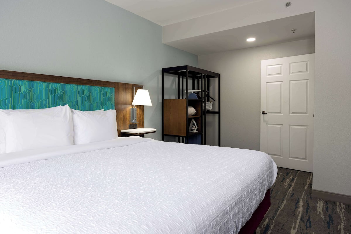 A hotel room with a large bed, a nightstand with a lamp, a wooden headboard, a built-in shelf, and a door.