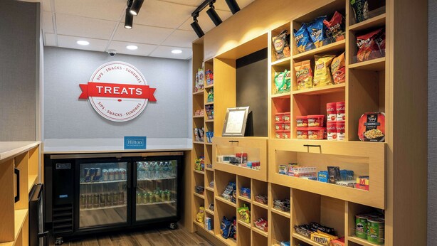 A hotel room with a minibar, a minibar fridge, and shelves stocked with snacks and beverages. - Hampton Inn & Suites Alpharetta Roswell (Alpharetta)