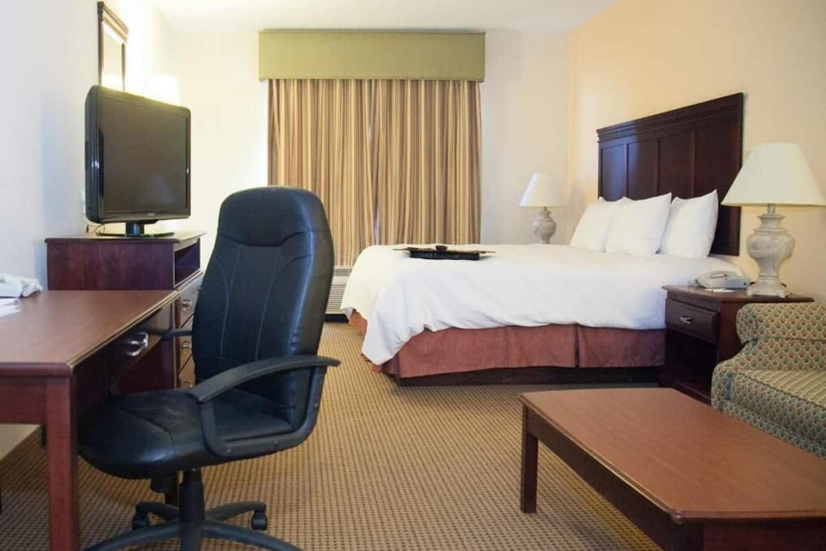 A hotel room with a bed, a desk with a television, a chair, a bedside table, and a lamp.