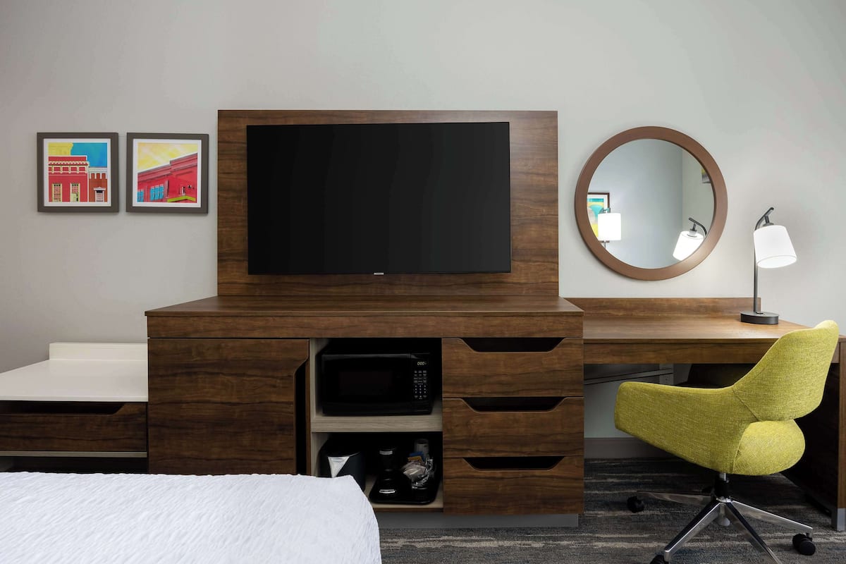 A hotel room with a wooden desk, a flat-screen TV, a round mirror, a yellow chair, and artwork on the wall.