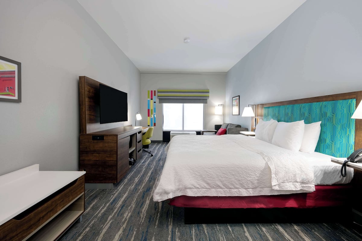 A hotel room with a large bed, a desk with a flat-screen TV, a chair, a desk lamp, and a window with striped curtains.