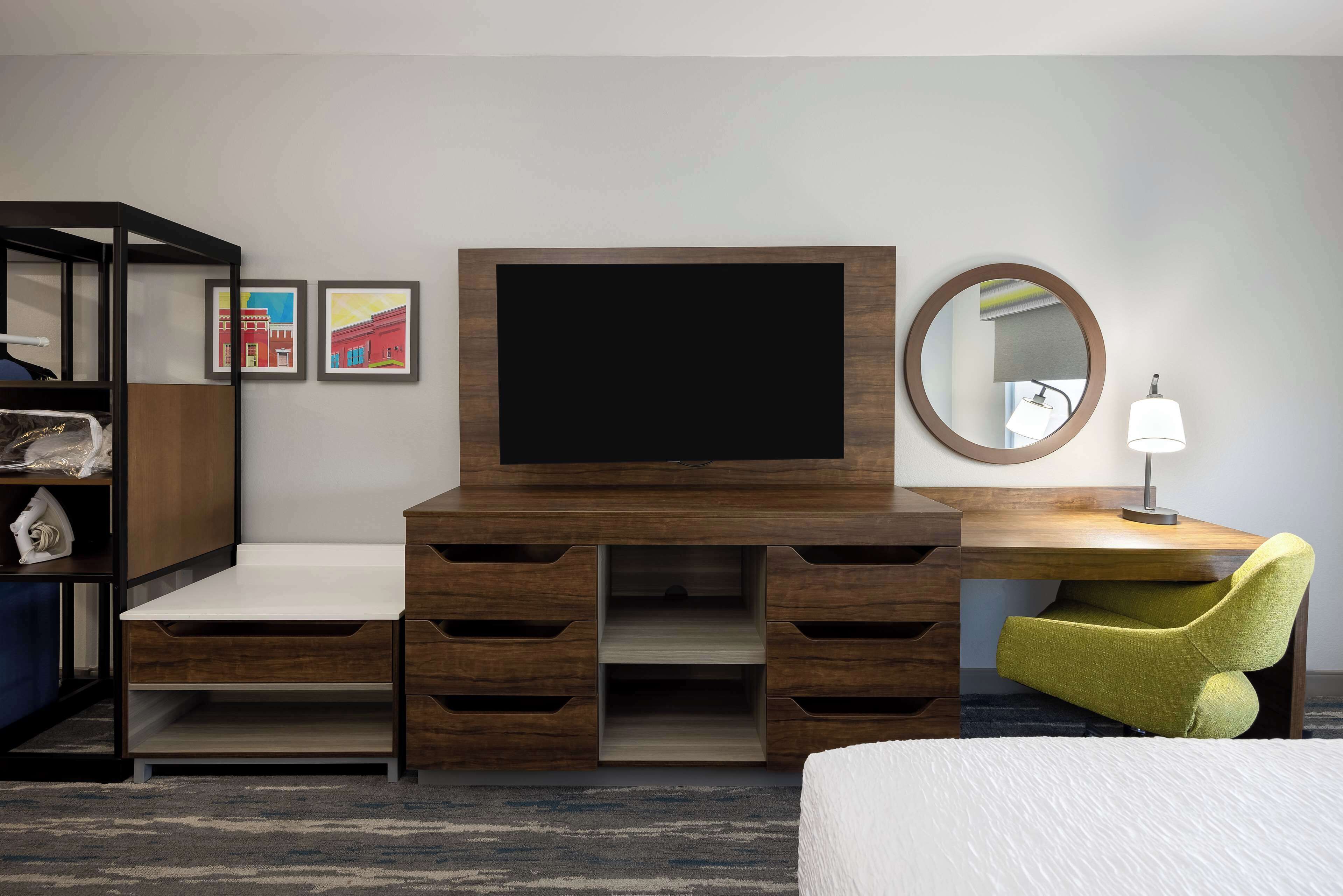 Una habitación de hotel moderna con un escritorio de madera, un televisor de pantalla plana, una silla verde, una cama y un pequeño estante con objetos decorativos.