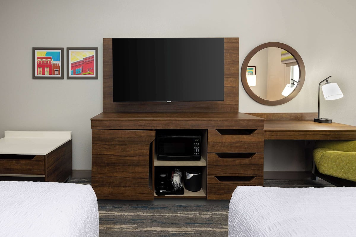A hotel room with a flat-screen TV, a round mirror, a desk with a lamp, and two beds.