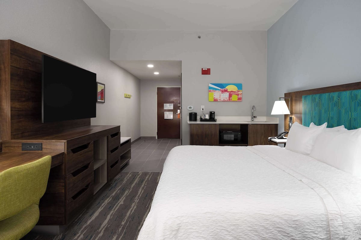 A hotel room with a large bed, a desk with a flat-screen TV, a desk lamp, a coffee maker, and a painting on the wall.