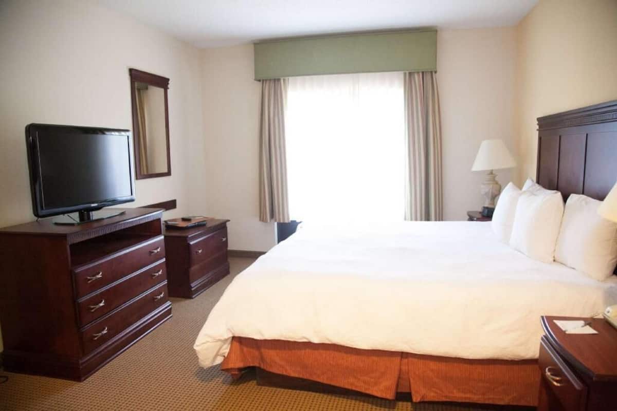A hotel room with a bed, a television, a dresser, a lamp, and a window with curtains.