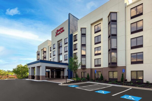 Un hotel moderno con un llamativo letrero de 'Hampton Inn Express Suites', un acceso cubierto y espacios de estacionamiento designados, incluyendo lugares para discapacitados.