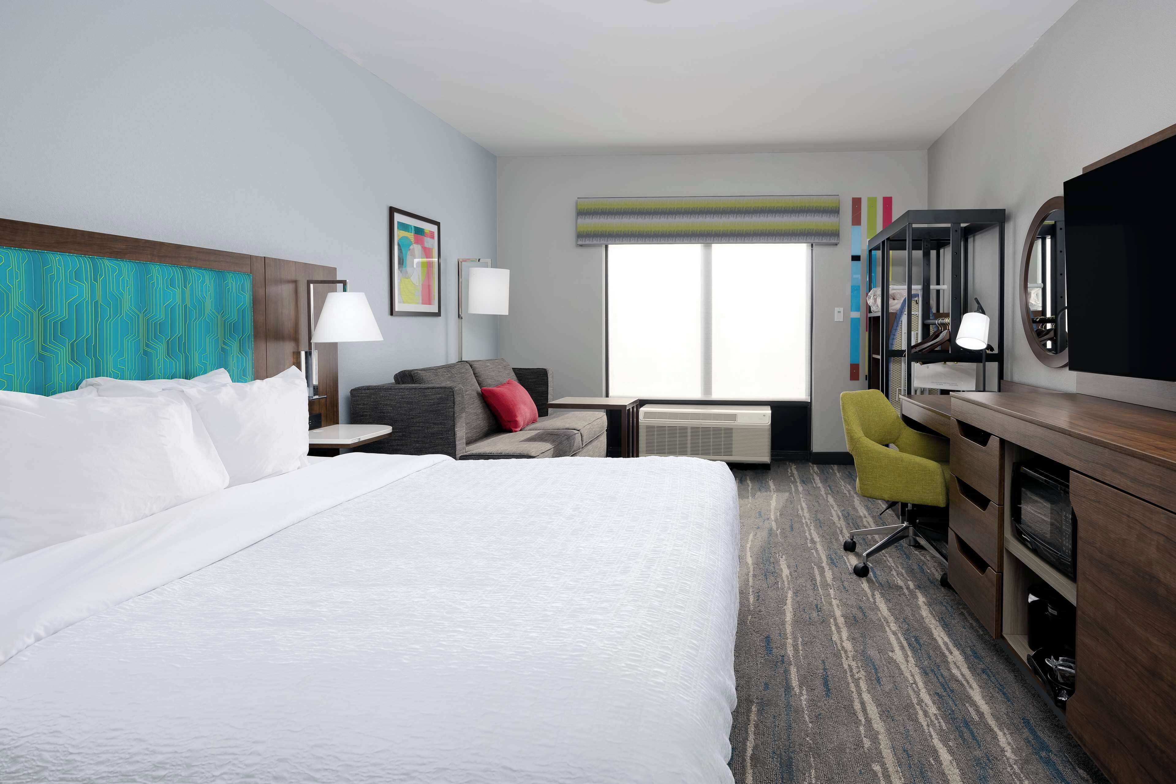 A hotel room with a large bed, a desk with a chair, a TV, a lamp, and a window with blinds.
