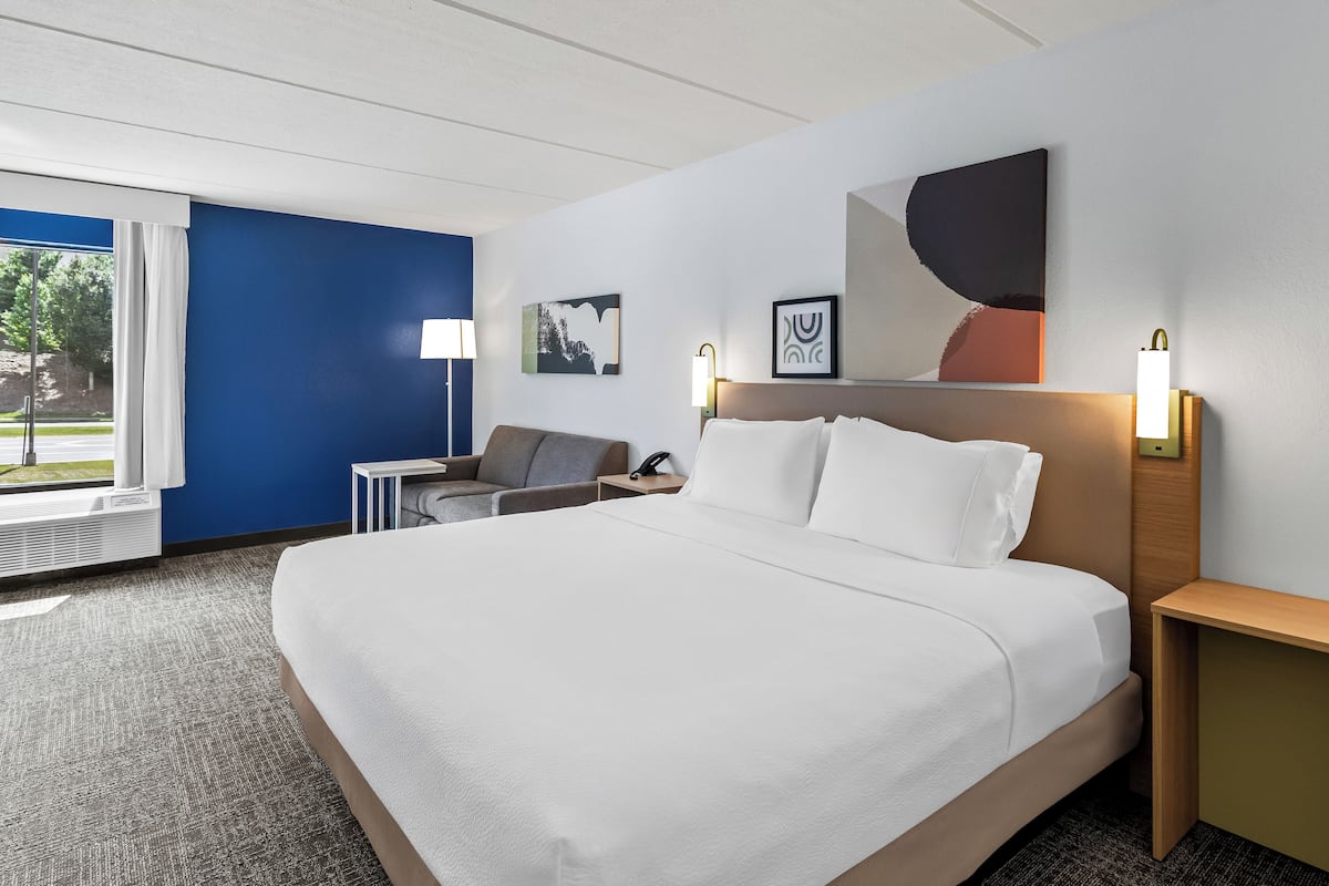 A hotel room with a large bed, a desk, and a chair. There is a window with curtains, a wall-mounted lamp, and a painting on the wall.