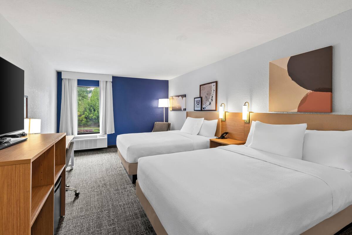 A hotel room with two beds, a desk, a TV, and a window with curtains.