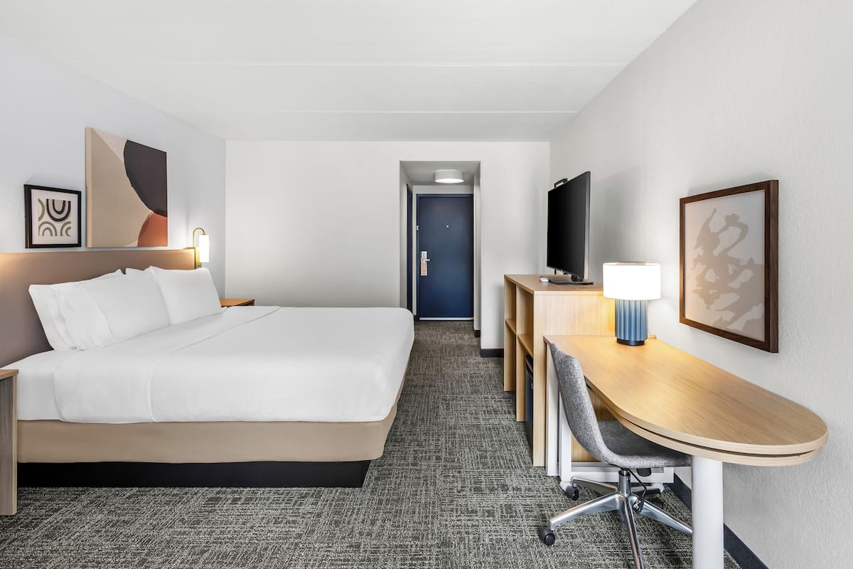 A hotel room with a bed, desk, chair, TV, and artwork.
