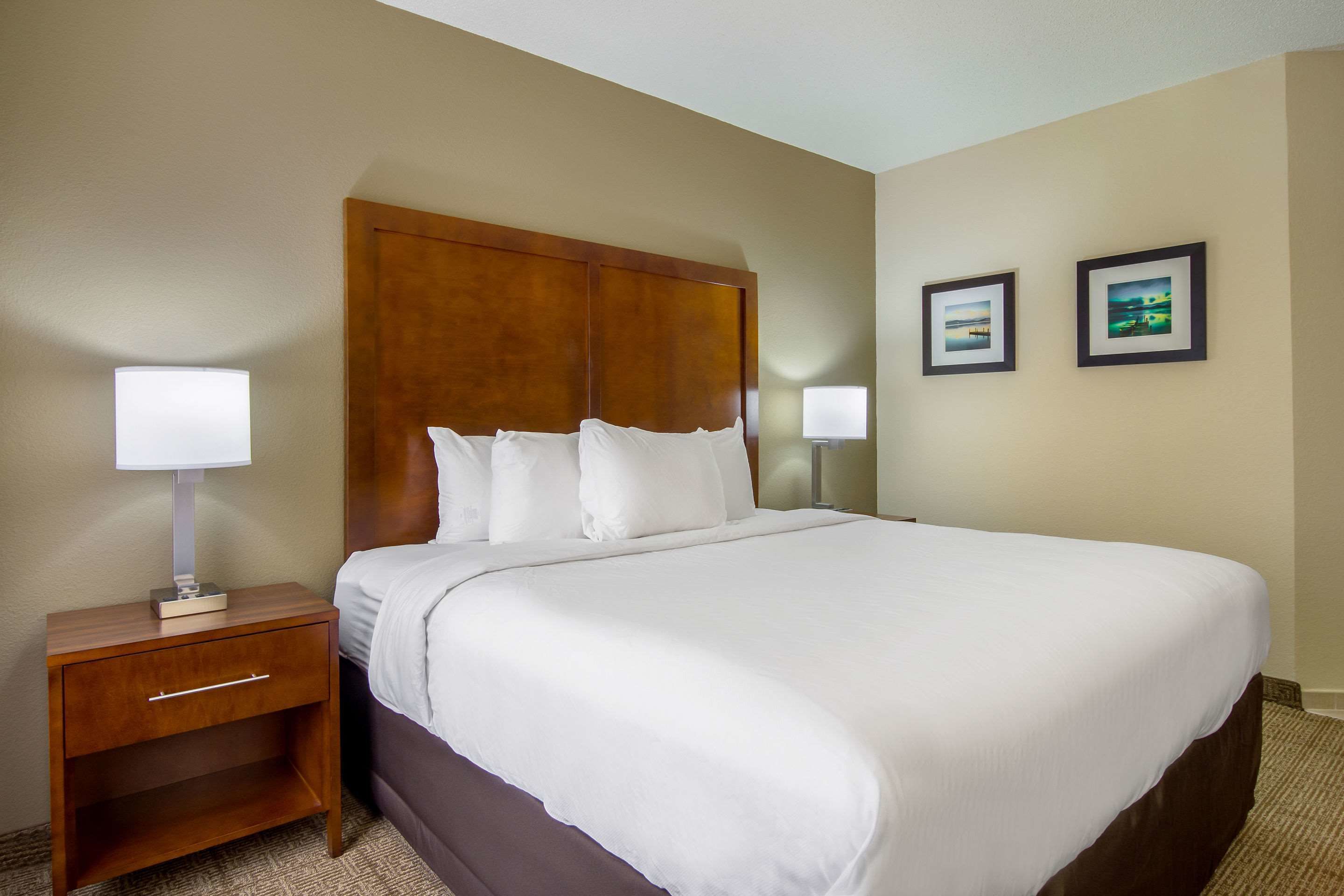 A hotel room with a large bed, a wooden headboard, a nightstand with a lamp, and two framed pictures on the wall.