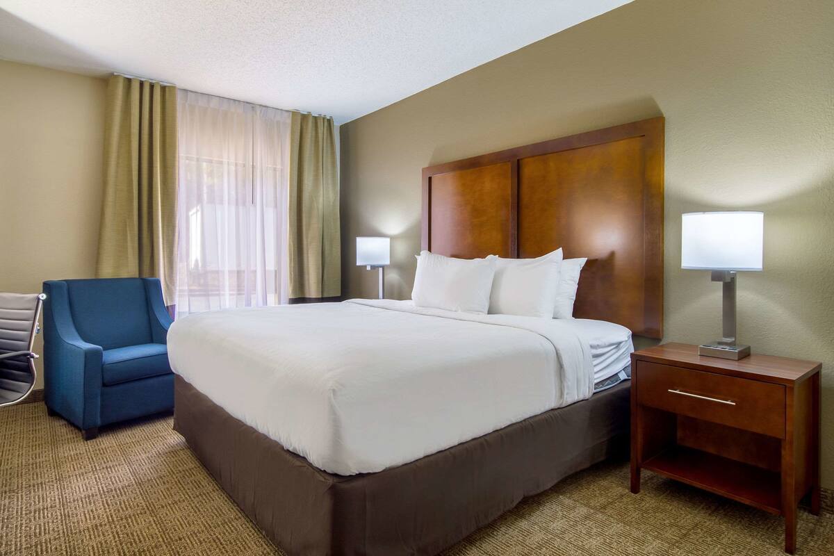 A hotel room with a large bed, two bedside lamps, a wooden nightstand, a blue armchair, and a window with sheer curtains.