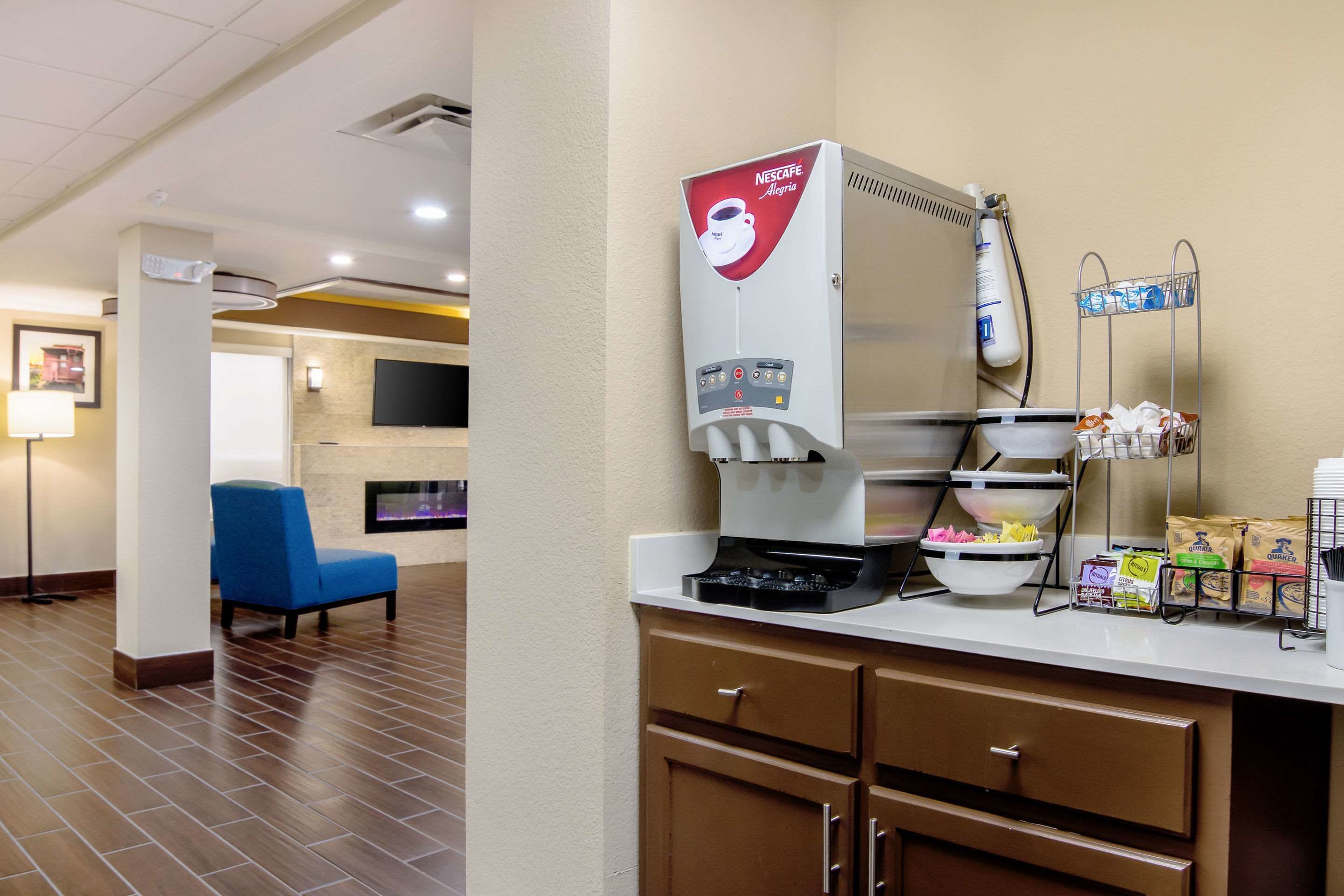 A Nescafé station with a coffee machine, a seating area with a blue chair, and a wall-mounted TV.
