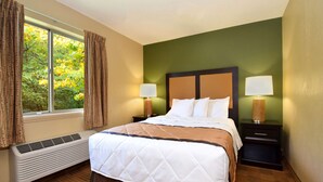 A hotel room with a bed, two bedside tables with lamps, a window with curtains, and a wall-mounted heater.