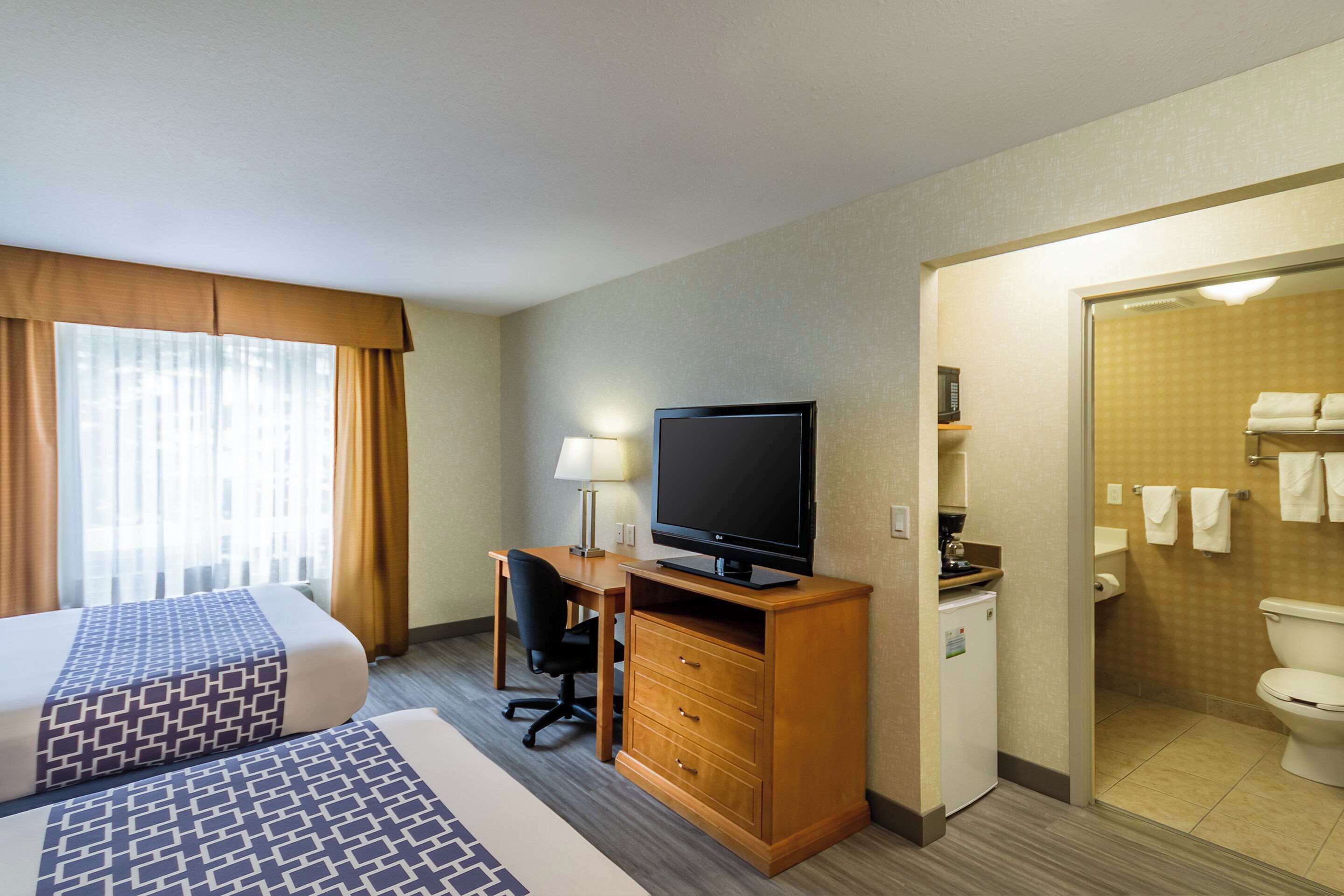 A hotel room with a bed, a desk with a television, a chair, a lamp, and a small refrigerator.