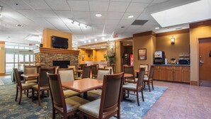 Un hall de hotel con varias mesas y sillas para comer, una chimenea de piedra, un televisor montado en la pared y una cocina visible a través de un vano abierto.