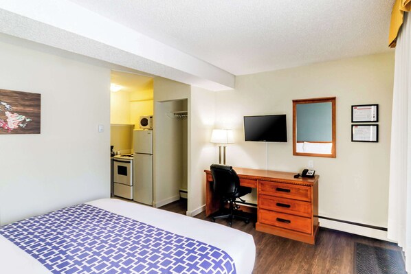 Una habitación de hotel con una cama, un escritorio con una silla, un televisor, un espejo y una pequeña kitchenette.