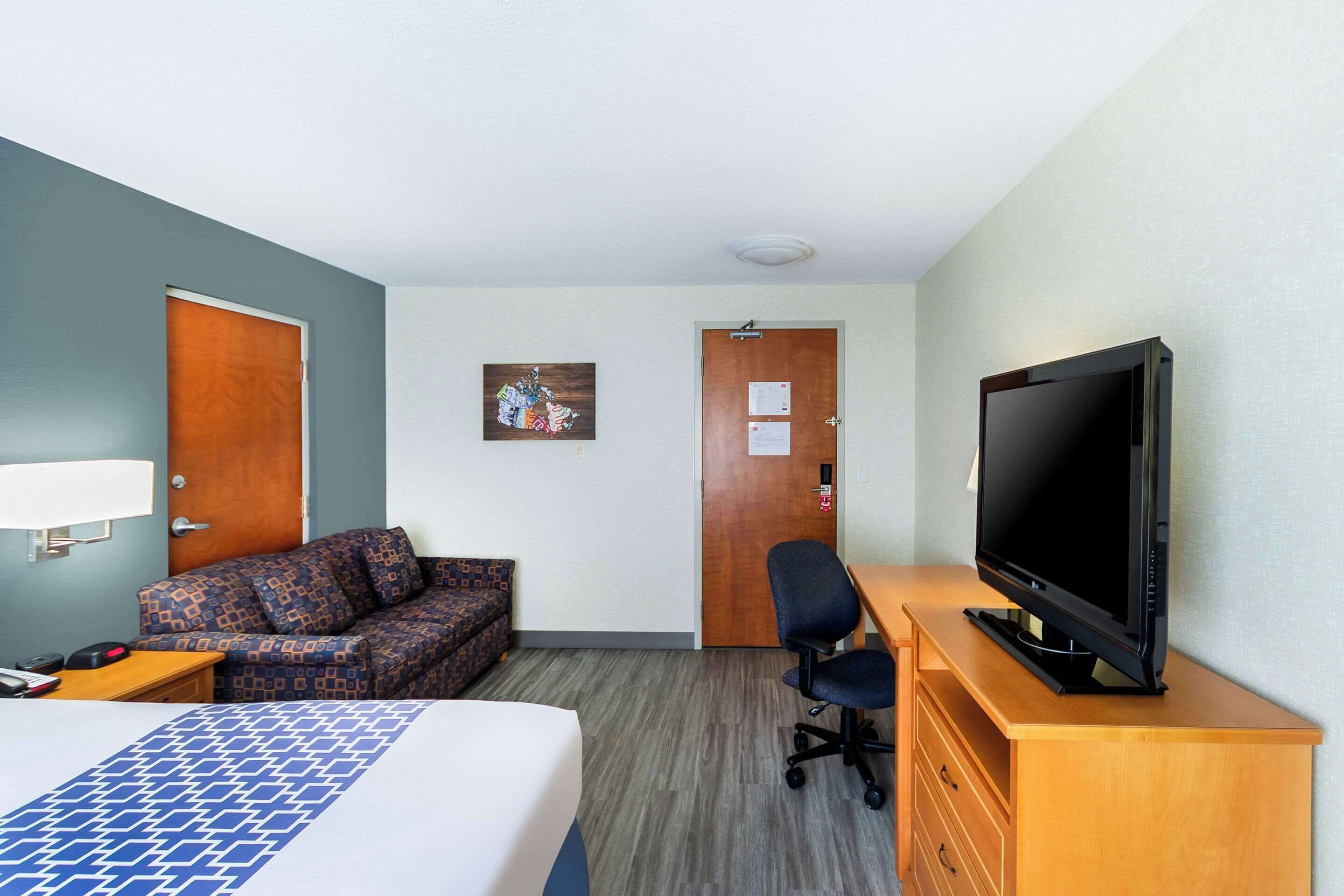 Una habitación de hotel con cama, sofá, escritorio con lámpara, televisor sobre un soporte de madera y puerta con tablón de anuncios.