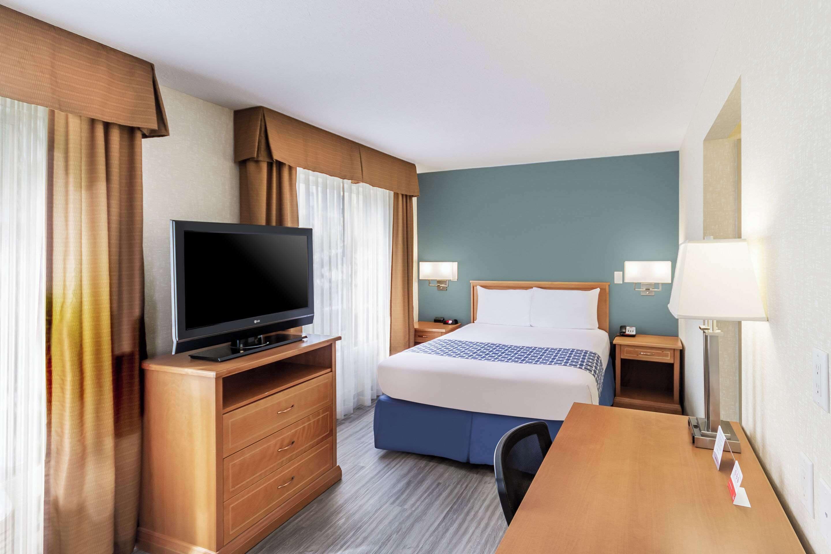 A hotel room with a bed, a TV on a wooden dresser, a desk, and two bedside lamps.
