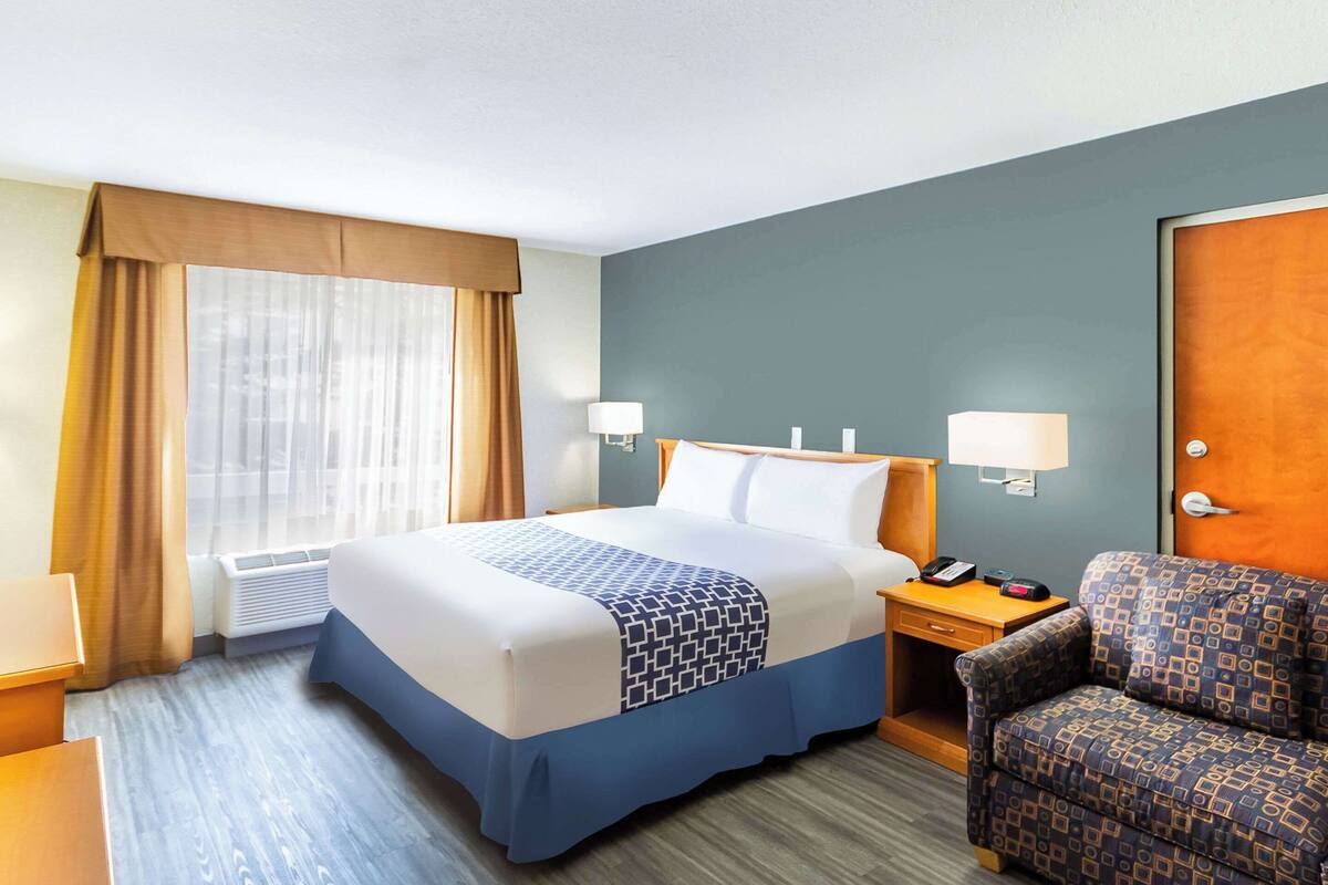 A hotel room with a bed, bedside table, lamp, chair, and a window with curtains.